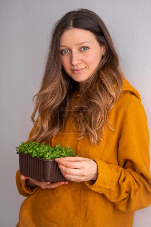 A contented woman in a mustard blouse holds a brown pot of dense microgreens, a nod to healthy eating and urban farming practices. Confident Woman in Mustard Sweater Holding a Pot of Fresh Microgreens