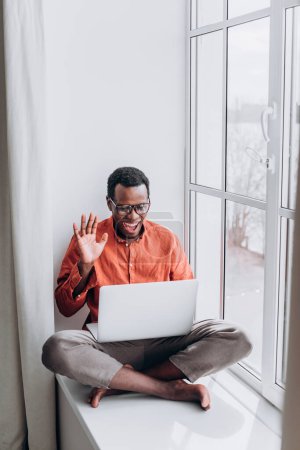 Homme afro-américain souriant Appel vidéo sur ordinateur portable par fenêtre. Un homme joyeux salue la webcam, s'engageant dans un appel vidéo sur son ordinateur portable avec la lumière naturelle coulant de la fenêtre à côté de lui