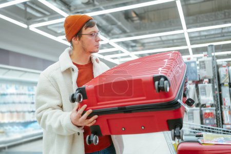 Mann trägt einen roten Hartschalenkoffer in einem Geschäft und erwägt Kauf. Nachdenklich betrachtet ein junger Mann in einem Geschäft einen roten Koffer und überlegt, ob er für seine Reisebedürfnisse geeignet ist.