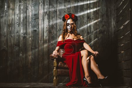 Eine bezaubernde Frau in Rot mit traditionellem Day of the Dead Make-up sitzt selbstbewusst da, ihre Pose verströmt einen geheimnisvollen Reiz. Geheimnisvolle Dame mit Tag der Toten Make-up