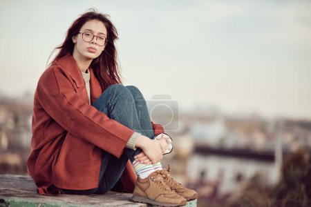 Foto de Chica adolescente tranquila pensativa en gafas con pelo largo y rojo en abrigo rojo se sienta con las rodillas dobladas y mira a la cámara, sobre el fondo del cielo y el paisaje urbano borroso. - Imagen libre de derechos