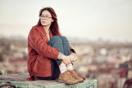 Foto de Chica adolescente pensativa en gafas con pelo largo y rojo en abrigo rojo se sienta con las rodillas dobladas y mira hacia otro lado, sobre el fondo del cielo y el paisaje urbano borroso. - Imagen libre de derechos