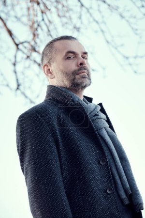 Photo for Portrait of a confident bearded middle-aged man in a blue coat and woollen scarf against the sky looking down at the camera. - Royalty Free Image