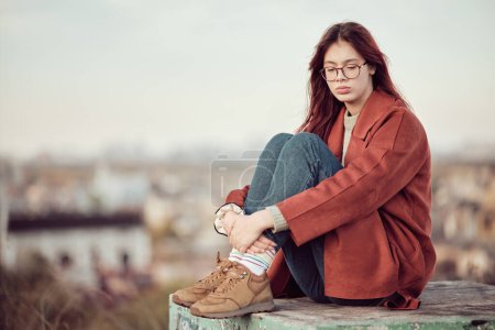 Foto de Chica adolescente pensativa en gafas con pelo largo y rojo en abrigo rojo se sienta con las rodillas dobladas sobre el fondo del cielo y paisaje urbano borroso. - Imagen libre de derechos