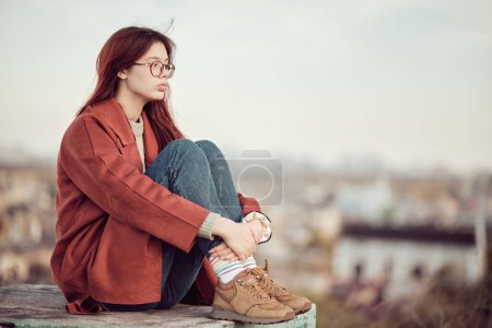 Foto de Chica adolescente pensativa en gafas con pelo largo y rojo en abrigo rojo se sienta con las rodillas dobladas y mira hacia otro lado, contra el fondo del cielo y el paisaje urbano borroso. - Imagen libre de derechos