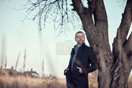 Man in a coat stands near a tree in a park in the countryside. Rest, slowing down, meditative mood.