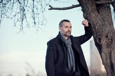 Portrait d'un homme d'âge moyen barbu en manteau bleu et écharpe de laine debout près d'un arbre.