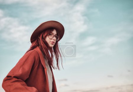 Teenage girl in red coat and hat stands against the sky and looks away.