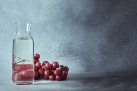 Téléchargez les photos : Carafe d'eau élégante avec raisins rouges sur fond gris texturé - en image libre de droit