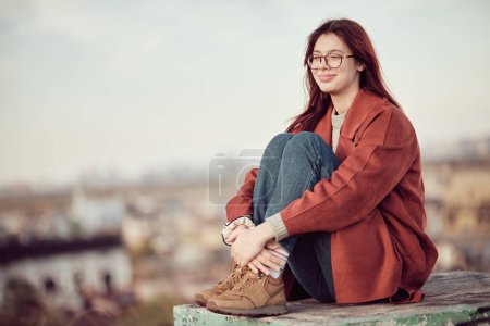Chica adolescente feliz y satisfecha en gafas con pelo largo y rojo en abrigo rojo se sienta con las rodillas dobladas y mira hacia otro lado, sobre el fondo del cielo y el paisaje urbano borroso.