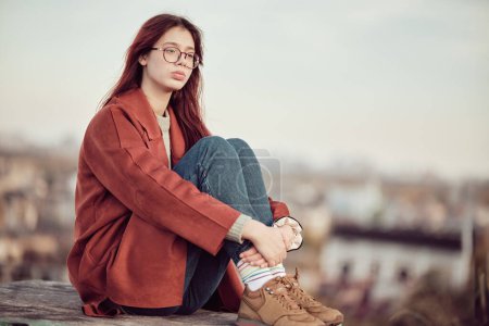 Chica adolescente pensativa en gafas con pelo largo y rojo en abrigo rojo se sienta con las rodillas dobladas y mira hacia otro lado, sobre el fondo del cielo y el paisaje urbano borroso.