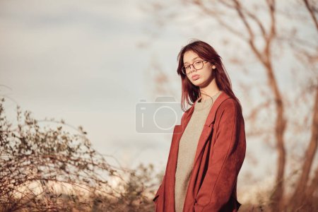 Adolescente confiante sérieuse avec de longs cheveux rouges en manteau rouge se tient dans le parc et regarde la caméra.