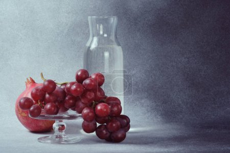 Water Carafe, Pomegranate and Red Grapes on Glass Stand, Textured Grey Background