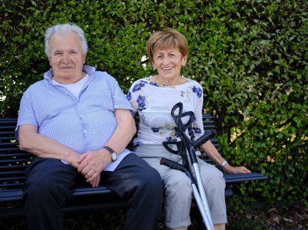Zwei ältere Rentner ruhen sich auf einer Bank im Stadtpark aus. Ältere Frau benutzt Krücken bei Gehproblemen, sie entspannt mit ihrem Mann auf einer Bank.