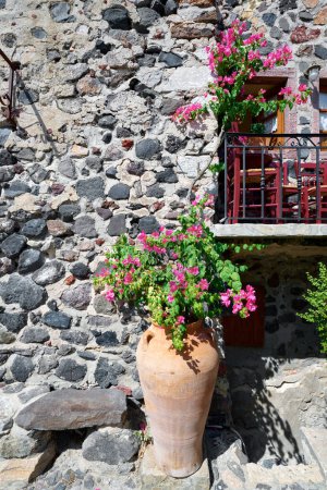 Schöne Vintage-Balkon mit bunten Blumen, Terrakottatopf, roten Stühlen und Natursteinmauer in einer engen Straße von Pyrgos Kallistis Dorf, Santorin. Griechische Inseln, Europäischer Urlaub.