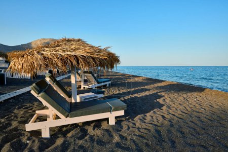 Sonnenschirme und Liegestühle aus Stroh am Strand von Perivolos mit schwarzem Sand und kleinen dunklen Kieselsteinen im Süden der Insel Santorin. Griechische Inseln, Santorin, Europaurlaub.
