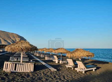 Sonnenschirme und Liegestühle aus Stroh am Strand von Perivolos mit schwarzem Sand und kleinen dunklen Kieselsteinen im Süden der Insel Santorin. Griechische Inseln, Santorin, Europaurlaub.