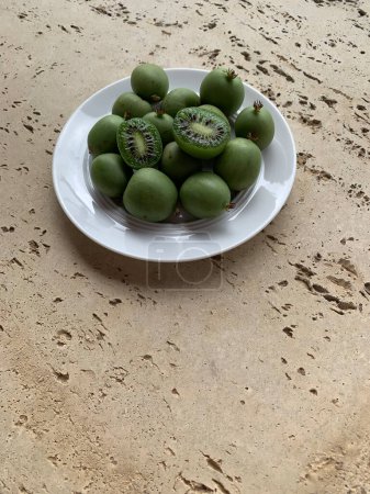 Téléchargez les photos : Assiette blanche avec kiwis ou mini kiwis verts entiers et demi sur fond de béton. Deux moitiés de kiwi coupées en deux dans des baies entières. Photo verticale. Place pour le texte. - en image libre de droit