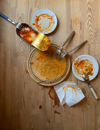 Photo for Dirty dishes and an oval tin can on a wooden background. View from above. Afternoon still life. Dirty saucers, forks, spoon, napkin, stains of tomato sauce on the table and board. - Royalty Free Image