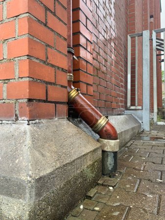 Ein braunes Abflussrohr, das vor dem Hintergrund einer braunen Ziegelmauer in Deutschland, der Stadt Ludwigsburg, in den Boden geht. Im Stil des alten Amerika. Seitenansicht.