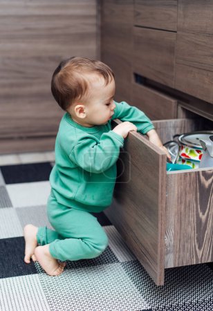 Téléchargez les photos : Petit garçon ouvrir les tiroirs de la cuisine pour jouer avec les ustensiles de cuisine à l'intérieur. Bébé explorant des objets de cuisine. Situation dangereuse - en image libre de droit