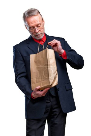 Foto de Senior man is looking at a paper bag receiving a present. Eco shopping. Man in suit on a white background - Imagen libre de derechos