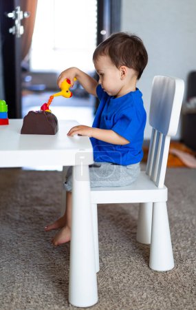 Téléchargez les photos : Jeune enfant absorbé dans le jeu à une table avec un jouet. Un petit garçon assis à une table jouant avec un jouet - en image libre de droit