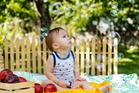 El bebé se sienta en una manta rodeada de burbujas. Un bebé feliz rodeado de burbujas de jabón flotantes en una manta colorida