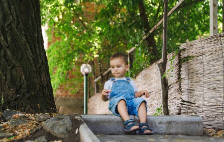 Téléchargez les photos : Un petit garçon assis sur une marche en ciment. Un enfant curieux contemplant les mystères de la vie - en image libre de droit