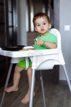 Téléchargez les photos : Un petit garçon assis sur une chaise haute mangeant. Un délicieux gâchis : l'aventure d'un petit garçon dans la chaise haute - en image libre de droit