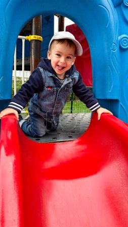 Téléchargez les photos : Petit garçon appréciant une diapositive colorée. Un petit garçon jouant sur un toboggan rouge et bleu. Un petit garçon jouant sur un toboggan rouge et bleu - en image libre de droit