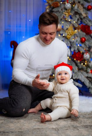 Photo for Capturing the magic of the holiday season, a man humbly kneels before a mesmerized baby, their hearts intertwined under the radiant glow of a sparkling Christmas tree. - Royalty Free Image