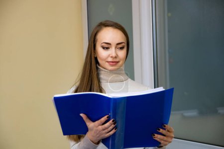 Photo for Woman Reading Blue Book in Front of Window. A woman sits in front of a window, engrossed in reading a blue book. - Royalty Free Image