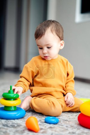 Téléchargez les photos : Un bébé joue avec une pile de blocs et un jouet. Le bébé porte une chemise jaune et est assis sur le sol - en image libre de droit