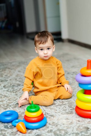 Téléchargez les photos : Un jeune garçon est assis sur le sol devant une pile de blocs colorés. Il porte une chemise et un pantalon jaunes. - en image libre de droit