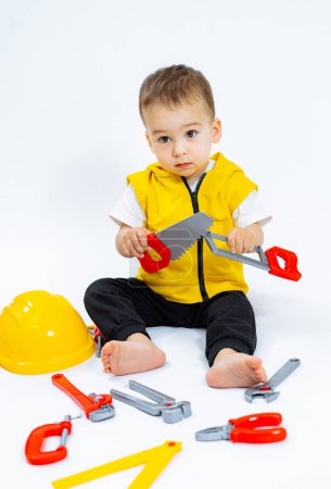 Téléchargez les photos : Un jeune garçon est assis sur le sol avec un marteau jouet et une scie à jouet. Il porte un gilet jaune et un chapeau jaune. - en image libre de droit