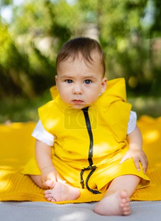 Téléchargez les photos : A baby is sitting on a blanket with a yellow jacket on. The baby has a blue eye and is looking at the camera - en image libre de droit