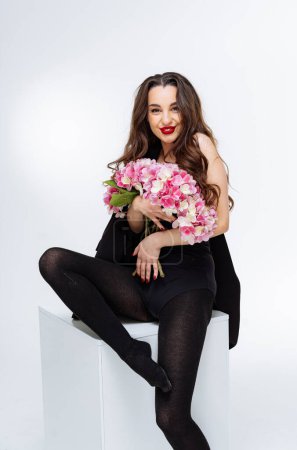 Téléchargez les photos : Une femme est assise sur une boîte blanche avec une fleur rose à la main. Elle porte des vêtements noirs et a du rouge à lèvres rouge. - en image libre de droit