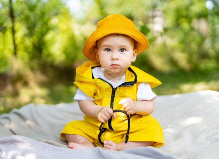 Téléchargez les photos : Un bébé portant un imperméable jaune et un chapeau est assis sur une couverture. Le bébé regarde la caméra avec un sourire sur le visage - en image libre de droit