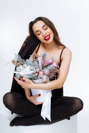 Téléchargez les photos : Une femme est assise sur une boîte blanche avec un bouquet de fleurs sur ses genoux. Elle porte une robe noire et un rouge à lèvres rouge. - en image libre de droit