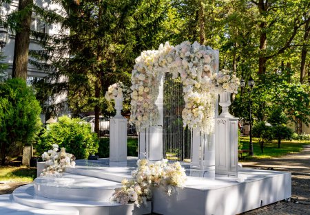 Une arche blanche avec des fleurs et une fontaine au milieu. La scène se déroule dans un parc avec des arbres et des buissons en arrière-plan