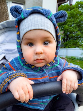 Téléchargez les photos : Un bébé portant un chapeau bleu et une veste rayée est assis sur une poussette. Le bébé a un grand sourire sur le visage et regarde la caméra - en image libre de droit