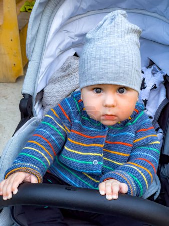 Téléchargez les photos : Un bébé portant un pull rayé et un chapeau est assis dans une poussette. Le bébé regarde la caméra avec une expression curieuse - en image libre de droit