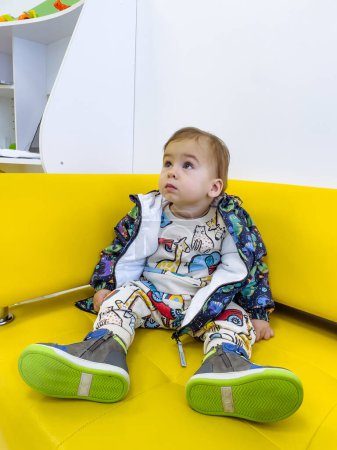 Téléchargez les photos : Un bébé est assis sur un canapé jaune portant une veste avec un dinosaure dessus. Le bébé regarde la caméra. - en image libre de droit
