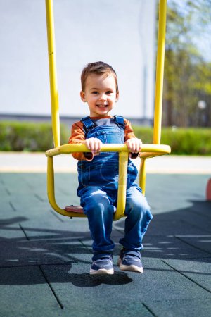 Téléchargez les photos : Un jeune garçon est assis sur une balançoire dans un parc. Il sourit et il s'amuse - en image libre de droit