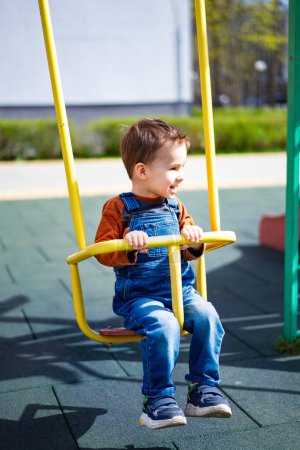 Téléchargez les photos : A young boy is sitting on a swing in a park. He is smiling and enjoying himself. The swing is yellow and is located near a green pole - en image libre de droit
