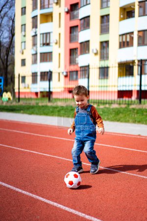 Téléchargez les photos : A young boy is playing with a soccer ball on a track. The boy is wearing a blue overalls and a brown shirt - en image libre de droit