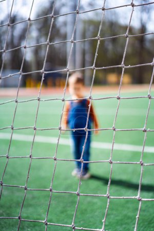 Téléchargez les photos : A young child stands in front of a soccer goal. The child is wearing a blue overalls and a red shirt. - en image libre de droit