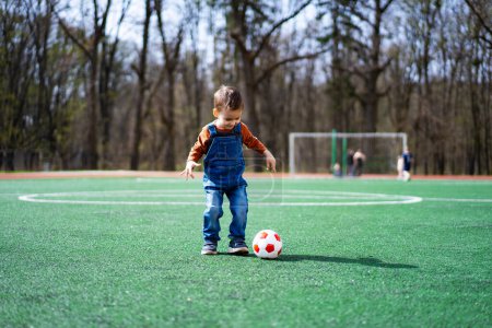 Téléchargez les photos : Un jeune garçon en salopette joue avec un ballon de football sur un terrain vert. Le garçon porte une chemise rouge et un jean bleu - en image libre de droit