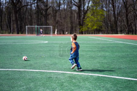 Téléchargez les photos : A young boy is playing soccer on a field. He is wearing a blue overalls and is holding a soccer ball - en image libre de droit